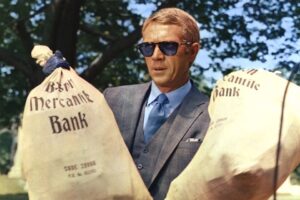 Thomas Crown-- actor Steve McQueen-- holding two very large money bags. 